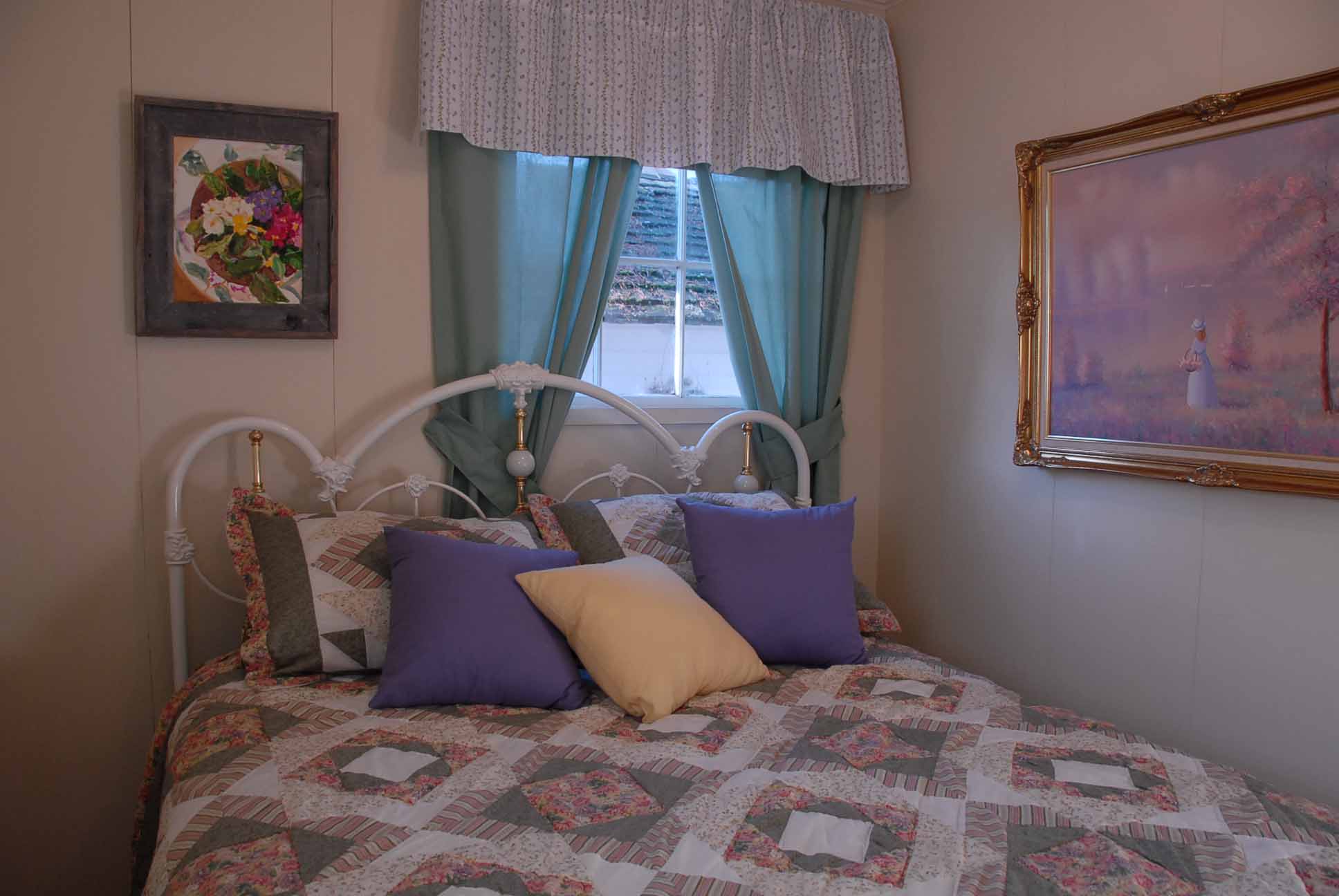The master bedroom, includes queen size bed with white iron Victorian headboard, a picture on the wall of a girl picking flowers by a lake and a picture of flowers